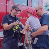 photo of firefighter in front of red fire engine letting toddler touch a firefighter helmet
