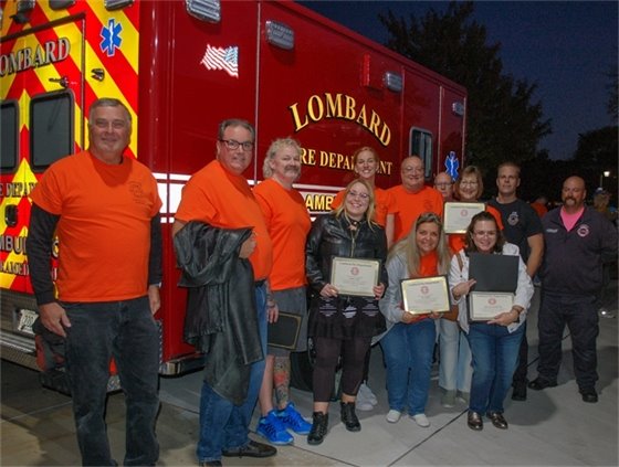 A group of 2025 Lombard Citizens Fire Academy graduates wearing orange shirts pose in front of a Lombard Fire Department ambulance. Several participants hold certificates of completion, joined by Lombard firefighters and instructors during the Community Open House event.