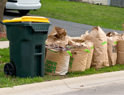 Green and yellow Waste Management toter alongside four paper yard waste bags on curb