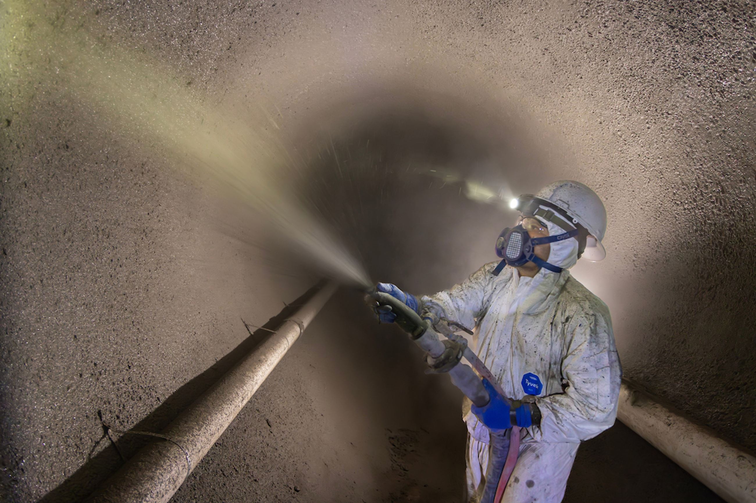 A worker wearing protective gear sprays geopolymer material along the inside of a large sewer pipe